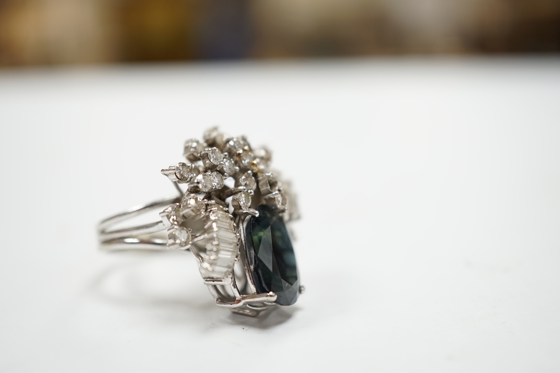 A mid to late 20th century 18k white metal, single stone oval cut sapphire and baguette and round cut diamond cluster set dress ring, size M/N, gross weight 10.3 grams.